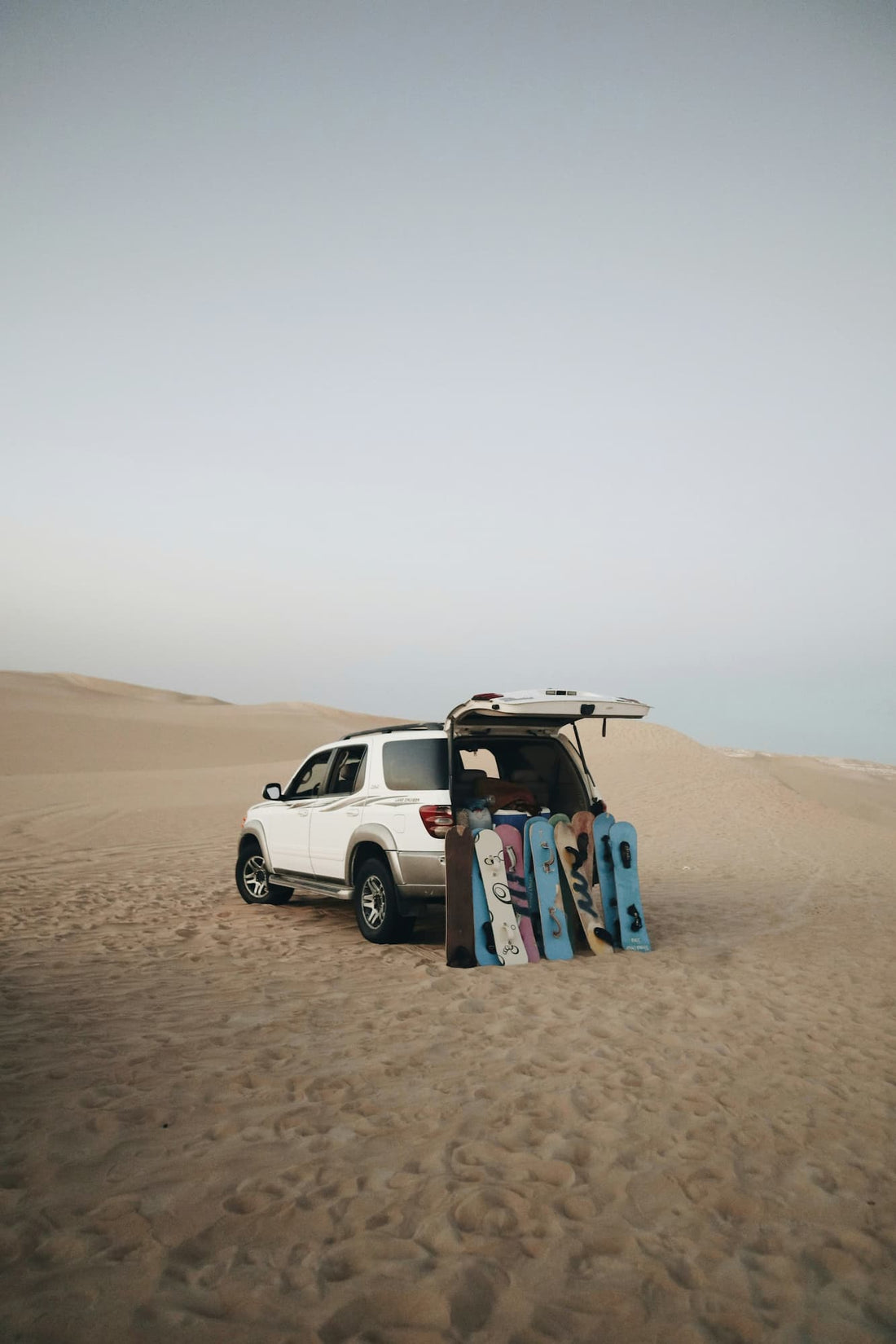 White SUV parked on sandy dunes with several sandboards propped against its open hatchback.