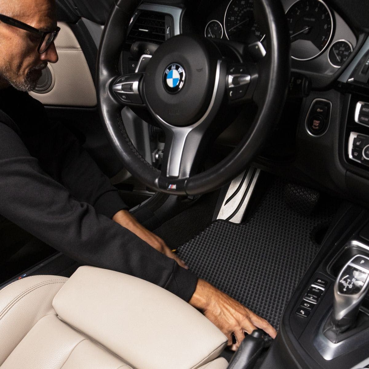 Driver installing EVA car mat in a BMW interior with black and beige leather finish.