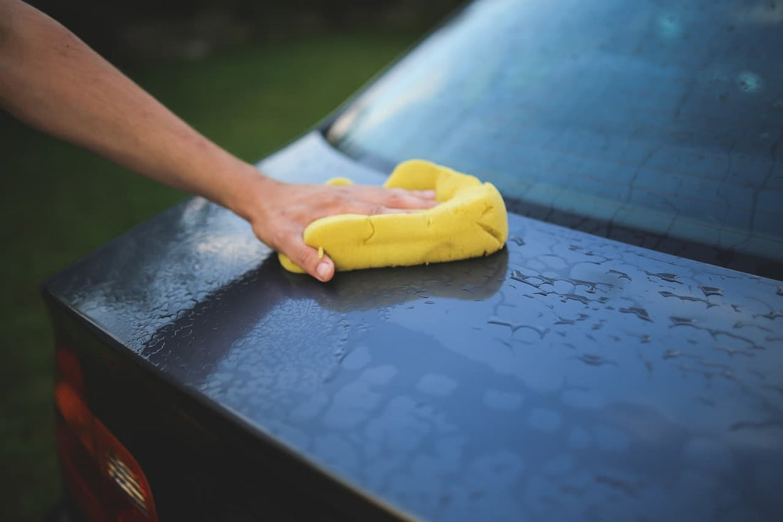 A person cleaning a wet car surface with a yellow sponge, removing water and dirt.