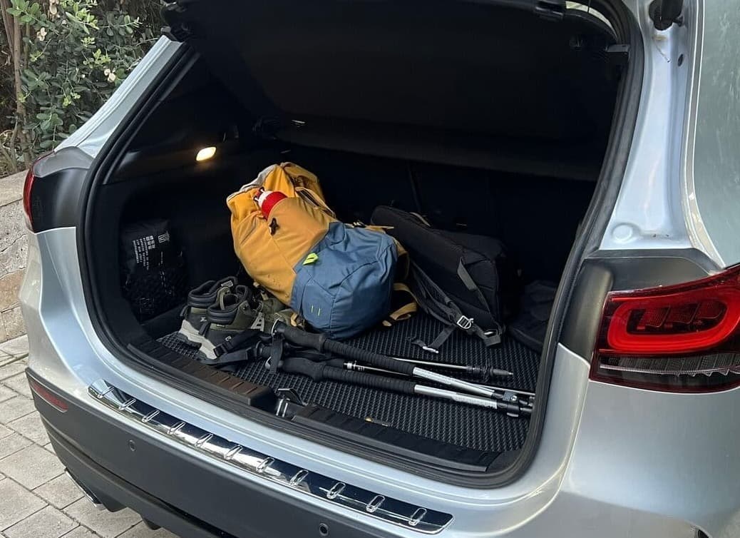 Open car trunk with hiking equipment, including backpacks, trekking poles, and shoes on a textured car mat.