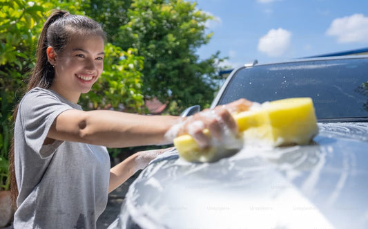 Smiling woman washing a car with a yellow sponge on a sunny day.