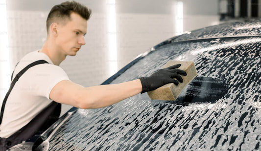 Man washing a car windshield with soap and sponge in a professional detailing shop.