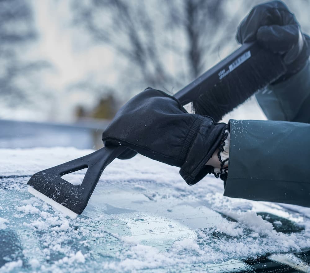 Person using snow scraper to remove ice from car windshield in winter.