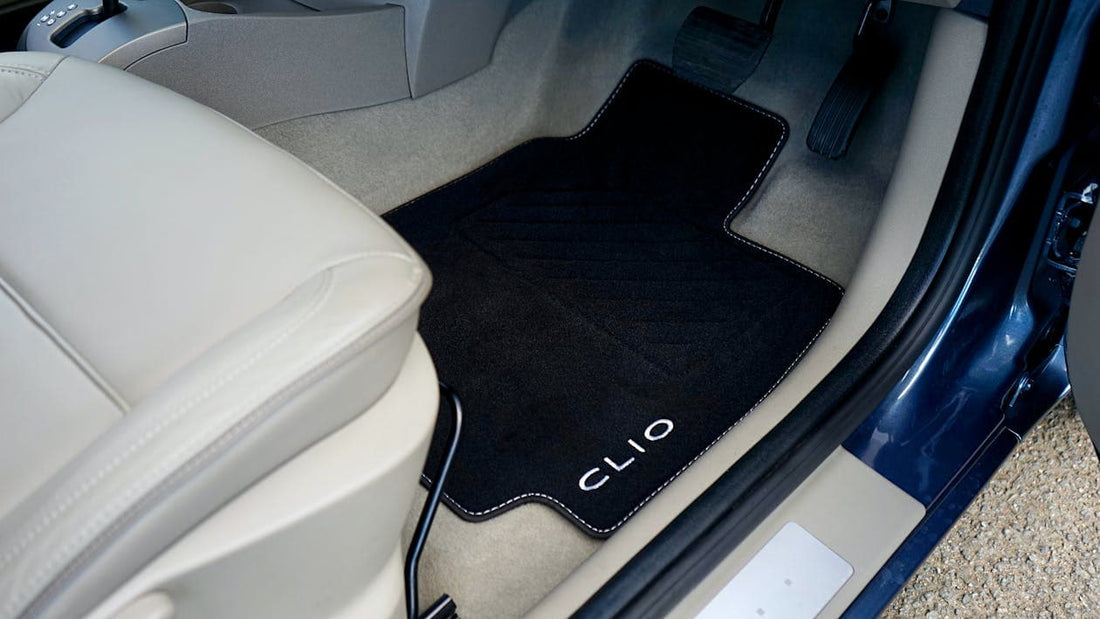 A custom black car mat with white stitching and "Clio" branding placed on the driver's side floor. 