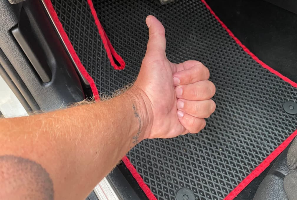 Thumbs-up hand gesture over black EVA car floor mat with red trim.