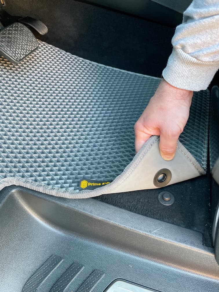 Hand lifting gray EVA car mat showing mounting detail near pedals.