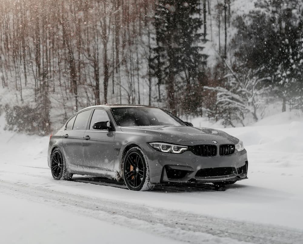Grey sedan driving on snowy winter road surrounded by trees.