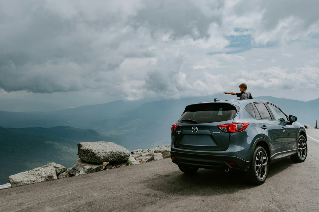 Blue Mazda SUV parked at a mountain overlook while a child stands through the sunroof pointing toward the distant scenery.