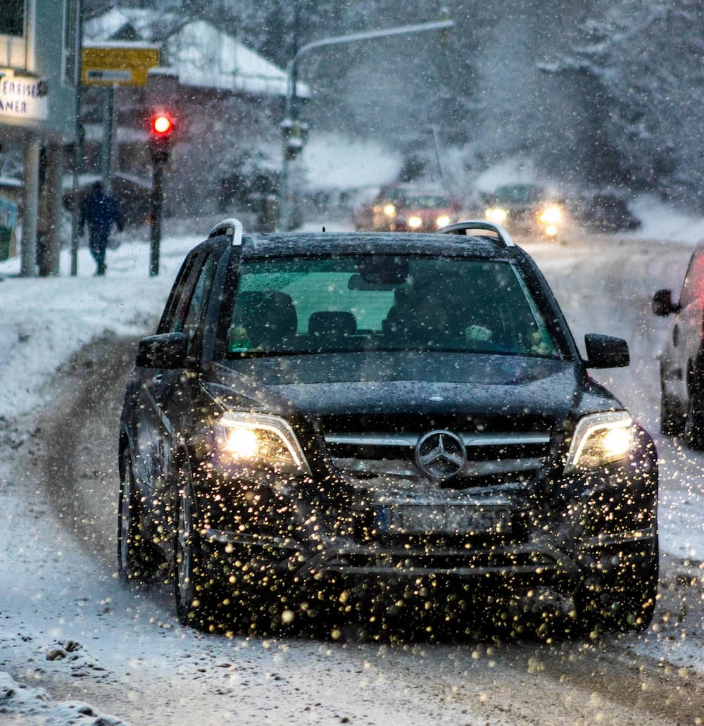Mercedes SUV driving through heavy snowfall on a snowy winter road with headlights on.