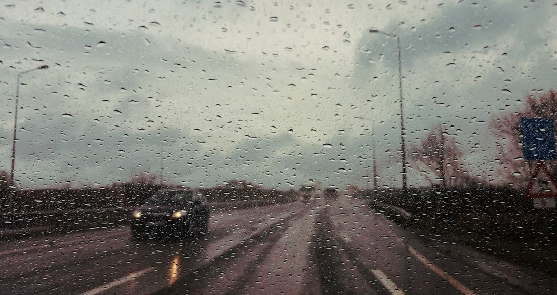 A rainy road view from a car windshield, with water droplets on the glass and a blurred vehicle ahead.