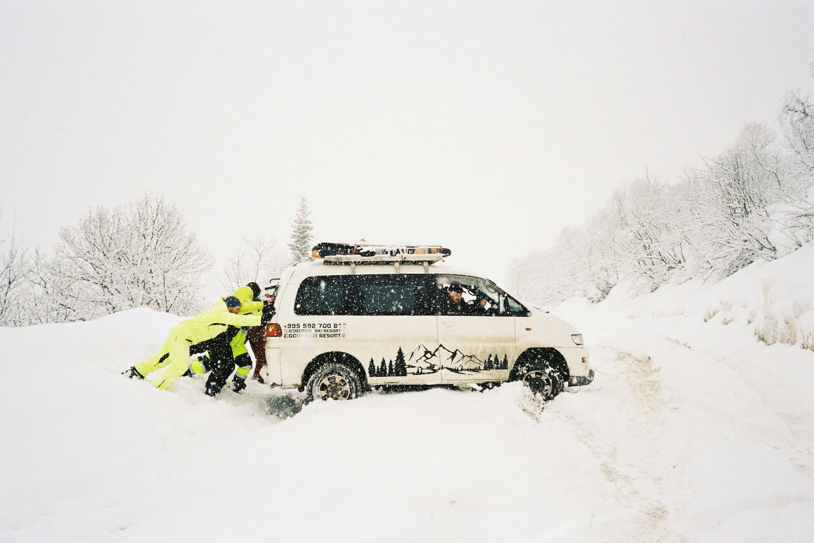 People push a van stuck in deep snow on a winter mountain road during heavy snowfall.