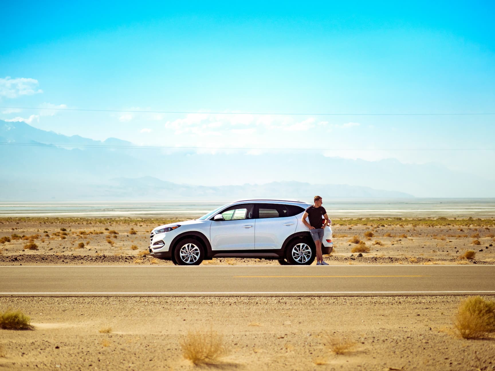 Man leaning on a white SUV parked on a desert road with mountains in the background.