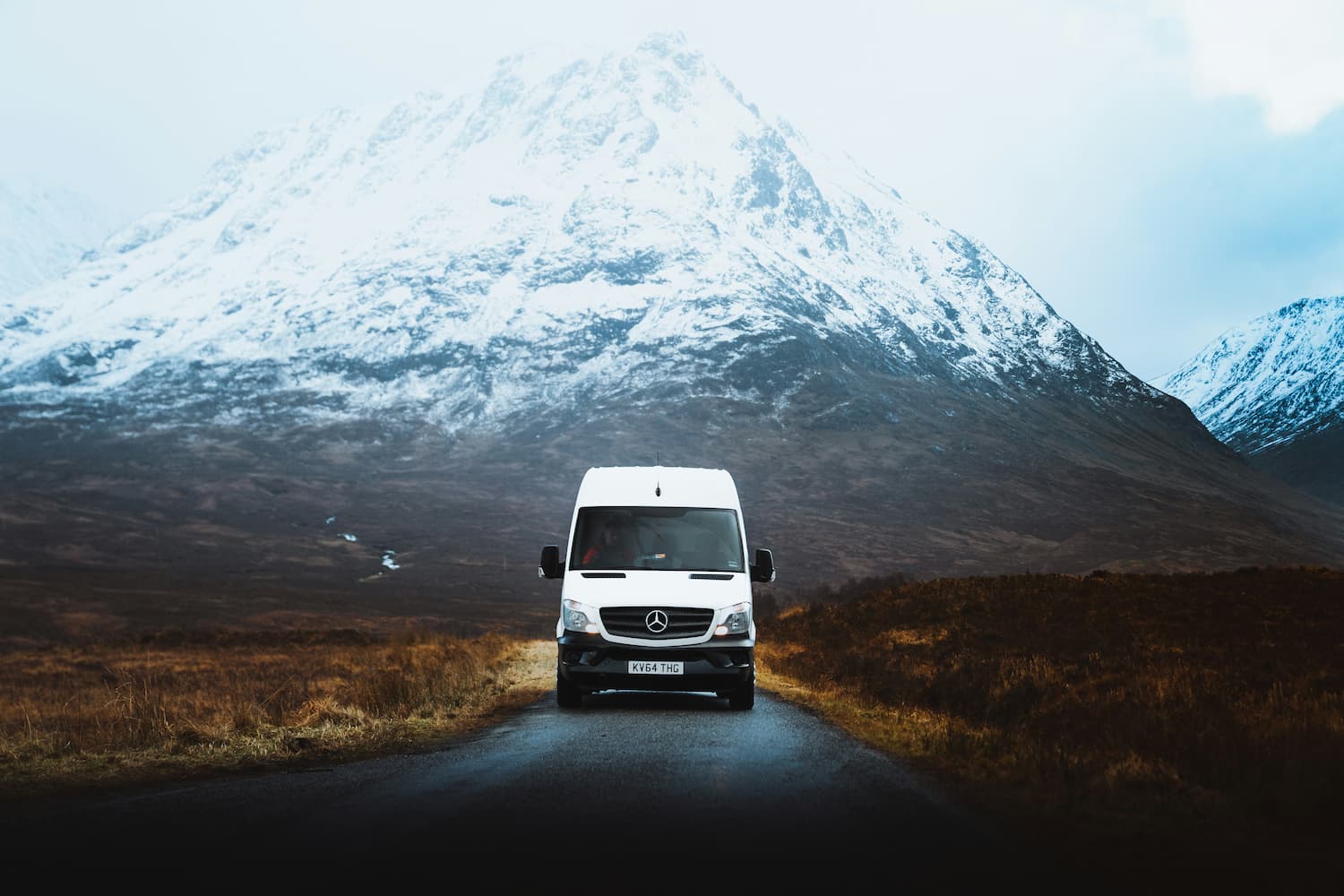White Mercedes van driving along remote mountain road surrounded by snow-covered peaks.