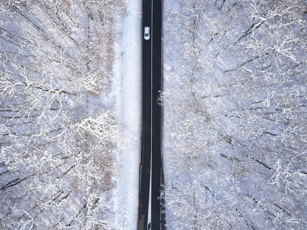 Aerial top view of a car driving on a straight winter road surrounded by snow-covered forest.