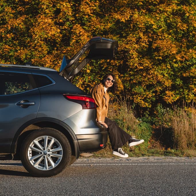 Woman sitting in the open car trunk enjoying a scenic autumn road view.