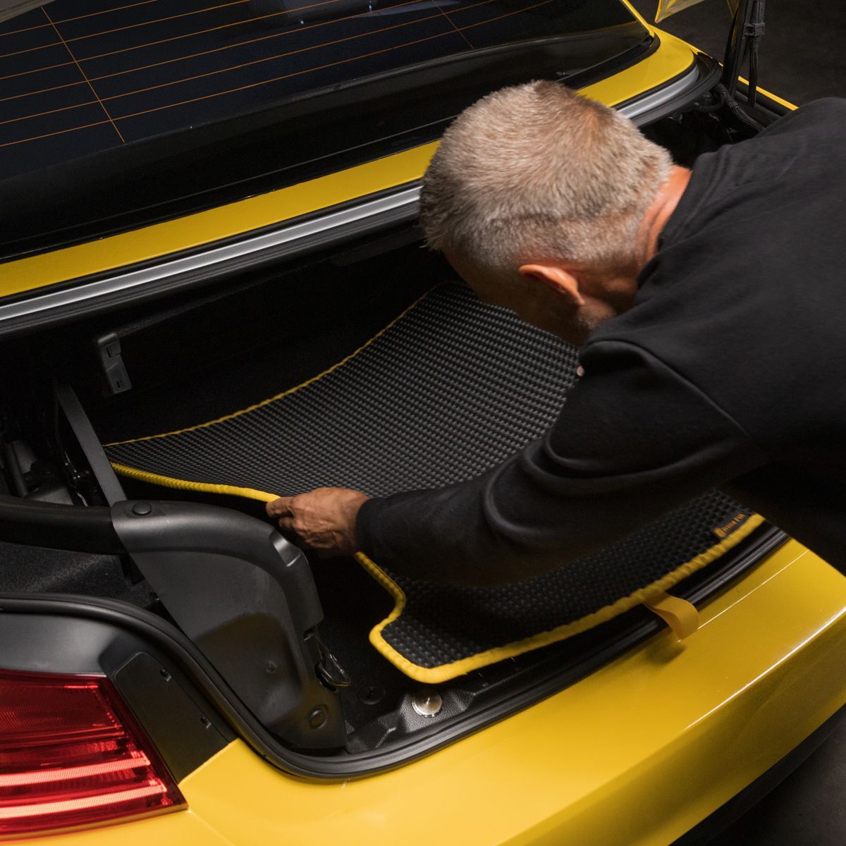 Custom-fit black and yellow trunk mat being installed in a BMW sports car.